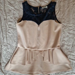 Beige ärmlös topp med spetsdetaljer - Säljer en elegant beige ärmlös topp från Kdk London med mörkblå spetsdetaljer upptill. Toppen har en peplumdesign som ger en fin siluett och en dragkedja i ryggen för enkel påklädning. Perfekt för en stilren look!