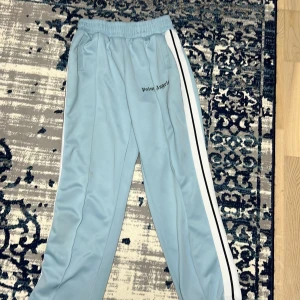 Sjuk Palm Angel äkta - Snygga ljusblå träningsbyxor från Palm Angels med vita och svarta ränder längs sidorna. Byxorna har en elastisk midja för extra komfort och en avslappnad passform. Perfekta för en sportig look.