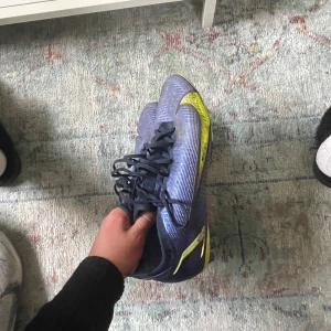 Blå/lila fotbollsskor från Nike - Snygga blå Nike Mercurial fotbollsskor med gula detaljer och snörning. Perfekta för fotbollsplanen med sin sportiga design och slitstarka material.