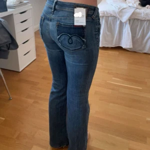 Blå jeans med broderade bakfickor - Snygga blå jeans med unika broderade detaljer på bakfickorna. De har en låg midja och en bootcut-stil som ger en avslappnad look. Perfekta för en casual outfit. Storlek 28:32 