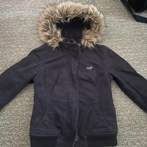 Hollister jacka - Säljer en jacka från Hollister i storlek M med fuskpäls på huvan och rutig insida. Jackan har dragkedja och knappar framtill samt en justerbar knytning i midjan. Perfekt för kyliga dagar!