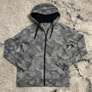 Grå camouflagemönstrad hoodie från Nike - Säljer en grå camouflagemönstrad hoodie från Nike med dragkedja och huva. Perfekt för en sportig look. Den har långa ärmar och en snygg Nike-logga på bröstet. Materialet är mjukt och bekvämt, idealiskt för kyligare dagar.