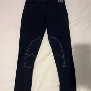 Svarta ridbyxor med kontrastsömmar - Snygga svarta ridbyxor med vita kontrastsömmar och förstärkningar vid knäna. Byxorna har en knappdetalj vid fickan och är designade för optimal komfort och rörelsefrihet vid ridning.