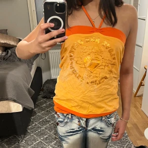 Orange halterneck topp - Trendig orange halterneck topp med textdetaljer framtill. Perfekt för en somrig look med sin livfulla färg och unika design. Passar bra till jeans eller kjol för en avslappnad stil.