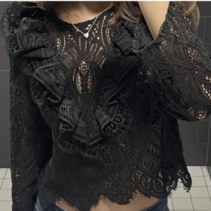 Svart spetsblus med volangdetaljer - Snygg svart spetsblus med vackra volangdetaljer och långärmad design. Perfekt för en elegant look med sitt genombrutna mönster och feminina stil. (Lånad bild) den är liten i storleken så skulle säga att den sitter med som en s/m