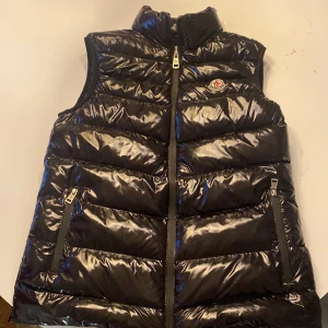Svart Moncler dunväst  - Snygg svart Moncler dunväst med glansig yta och dragkedja framtill. Västen har två praktiska fickor med dragkedjor och en hög krage för extra värme. Perfekt för kyliga dagar. En liten skada i insidan av västen. Priset kan diskuteras.