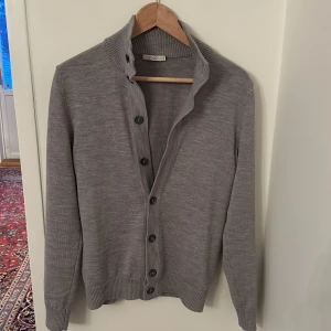 Grå kofta från Suitsupply - Snygg grå kofta från Suitsupply i merinoull. Den har en klassisk design med knappar framtill och ribbade detaljer vid krage, ärmslut och nederkant. Har ett litet håll på armen bild 4.  Säljer för den är för liten på mig.