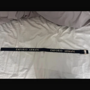 Svart bälte från Emporio Armani - Snyggt svart bälte från Emporio Armani med guldspänne och tydlig logotyp i guld. Perfekt för att ge en lyxig touch till din outfit. Bältet har en klassisk design med justerbar längd. Aldrig använd och köpt i Milano 2019