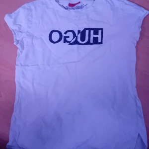 Vit t-shirt från Hugo - Säljer en vit t-shirt från Hugo med ett stort, spegelvänt tryck i blått på framsidan. T-shirten har korta ärmar och en klassisk passform. Perfekt för en avslappnad stil. XS