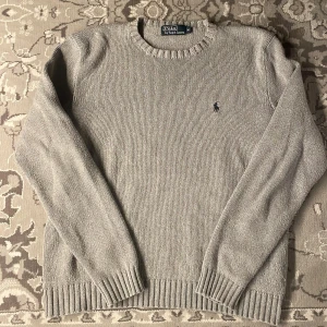 Grå stickad tröja från Polo Ralph Lauren - Säljer en stilren grå stickad tröja från Polo Ralph Lauren med det klassiska broderade logotypen på bröstet. Tröjan har en rund halsringning och ribbade muddar vid ärmslut och nederkant. Perfekt för en avslappnad och elegant look.