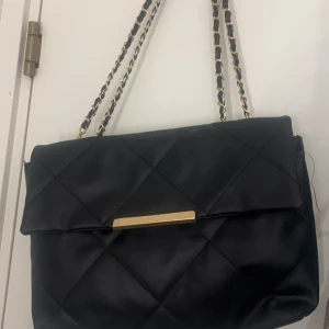 Svart quiltad axelväska med kedjedetaljer - Snygg svart axelväska med quiltat mönster och eleganta kedjedetaljer på axelremmen. Väskan har en stilren design med en guldfärgad detalj på framsidan. Perfekt för att ge en touch av lyx till din outfit.