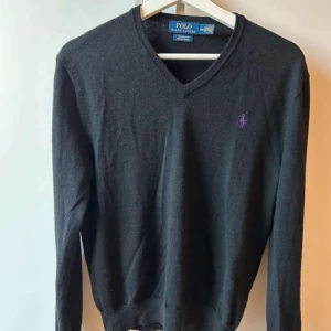 Svart tröja från Polo Ralph Lauren - Storlek M, Snygg svart tröja från Polo Ralph Lauren med lila broderad logga på bröstet. Tröjan har en v-ringad hals och långa ärmar, perfekt för en stilren look.