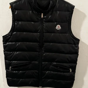 Moncler Gui Väst - Snygg svart dunväst från Moncler med dragkedja och två fickor framtill. Perfekt för kyligare dagar och har en stilren design med Moncler-logga på bröstet.