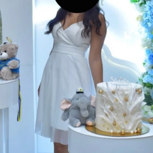 Studentklänning, vit klänning med omlott och glitterdetaljer - Elegant vit klänning med omlott-design och subtila glitterdetaljer. Klänningen har en v-ringad urringning och är ärmlös, vilket ger en stilren och festlig look.