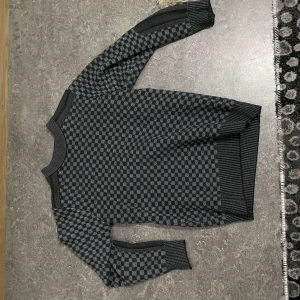 Grå och svart rutig tröja - Snygg grå och svart rutig tröja med långa ärmar och ribbade muddar. Perfekt för kyligare dagar med sitt stilrena mönster.