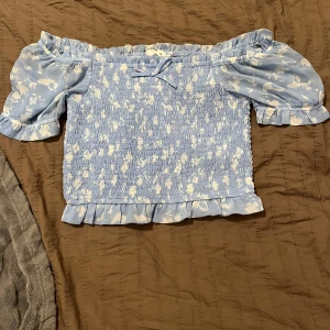 Blå offshoulder topp med blommönster - Charmig blå offshoulder topp med vita blommor och volangdetaljer. Toppen har en söt rosett framtill och puffiga kortärmar. Perfekt för en somrig look!😊