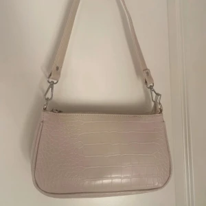 Beige axelväska från Gina Tricot - Snygg beige axelväska i syntet från Gina Tricot med krokodilpräglat mönster. Väskan har en praktisk dragkedja och en justerbar axelrem. Perfekt för att bära dina nödvändigheter med stil.