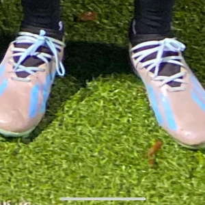 Messi fotbollsskor  - Snygga fotbollsskor med ljusblå och beige färgkombination. Skorna har snörning och är perfekta för fotbollsplanen. De har en modern design med kontrasterande blå ränder.