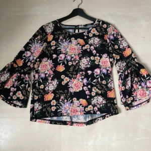 Blommig blus med vida ärmar - Säljer en svart blus med färgglatt blommönster i rosa, lila och orange. Blusen har vida trekvartsärmar och en rund halsringning. Perfekt för en färgglad look!