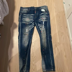 Blå jeans från Dsquared2 - Snygga blå jeans från Dsquared2 med unika färgstänk och slitna detaljer. Jeansen har en straight passform och är tillverkade i Italien. Perfekta för dig som vill ha en cool och avslappnad stil.