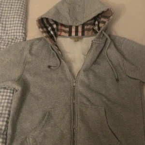 Grå huvtröja från Burberry - Snygg grå huvtröja från Burberry med klassiskt rutigt mönster i fodret. Tröjan har dragkedja och en bekväm huva. Perfekt för en stilren och avslappnad look.