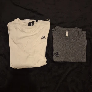 Två t-shirts från Adidas - Säljer två snygga t-shirts från Adidas. En vit och en grå, båda med korta ärmar och den klassiska loggan på bröstet. Perfekta för träning eller vardagsbruk.
