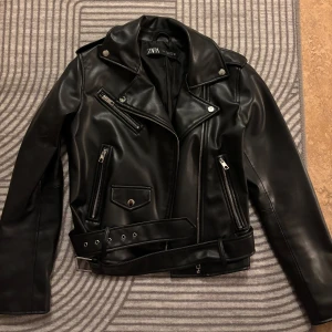 Svart skinnjacka från Zara - Snygg svart skinnjacka från Zara med dragkedjor och bälte i midjan. Jackan har en klassisk bikerstil med flera fickor och detaljer i silver. Perfekt för en cool look!