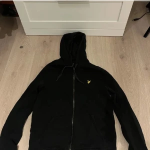 Svart hoodie med dragkedja - Säljer en svart hoodie med dragkedja och huva. Den har ett litet gult broderat emblem på bröstet. Dragkedjan är lite sönder kolla bild 3 men annars inga defekter!