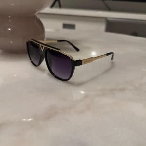 Solglasögon från Louis Vuitton - Snygga solglasögon från Louis Vuitton med svart och guld färgkombination. De har en elegant design med en V-formad detalj på framsidan och märkets logotyp på skalmarna. Perfekta för en stilren look.