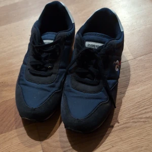 Blå sneakers från Le Coq Sportif - Snygga blå sneakers från Le Coq Sportif jr storlek 35  med svarta detaljer och vit logga på sidan. Skorna har en röd och vit sula samt snörning för en bekväm passform. Perfekta för en sportig look.