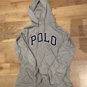 Grå hoodie från Polo Ralph Lauren - Säljer en grå hoodie från Polo Ralph Lauren med stor POLO-text i mörkblått på framsidan. Perfekt för en avslappnad stil. Modellen är 171 cm och 65kg. OBS! Tröjan har en pytteliten defekt i nederdelen som knappt syns.