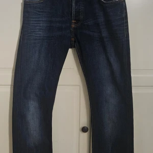 Mörkblå jeans från Nudie Jeans - Snygga mörkblå jeans från Nudie Jeans med klassisk femficksdesign och orange kontrastsömmar. Byxorna har knappar i gylfen och en läderpatch med logga baktill. Perfekta för en stilren look.                                   Ursprungliga priset är 1600 kr/ nytt pris 499     