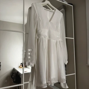 Vit klänning med knytband (studentklänning) - Elegant vit klänning med v-ringning och långa ärmar. Den har ett knytband i ryggen som ger en fin detalj och passform. Perfekt för en stilren look. Endast använt 1 gång på min student. 