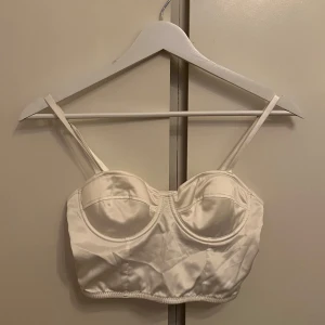 Vit ”satin” topp - Elegant vit korsett i satin med justerbara axelband och hakknäppning bak. Perfekt för en stilren look. I ett mycket bra skick!