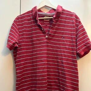 Röd randig pikétröja från Tommy Hilfiger - Snygg röd pikétröja från Tommy Hilfiger med vita ränder och klassisk krage. Tröjan har korta ärmar och knappar framtill. Perfekt för en avslappnad stil.