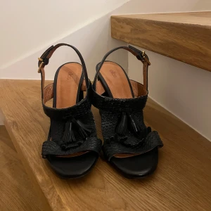 Svarta sandaler med klack och tofsdetalj - Snygga svarta sandaler med klack och dekorativa tofsar framtill. Sandalerna har en justerbar rem runt ankeln och en elegant fyrkantig tå. Perfekta för en stilren look.