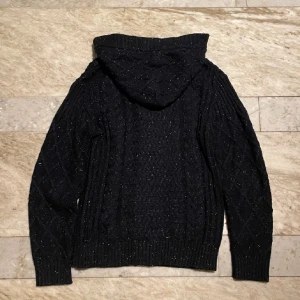 Svart stickad hoodie med knappar - Mysig svart stickad hoodie med knappar framtill. Tröjan har ett snyggt mönster och en praktisk huva. Perfekt för kyliga dagar.