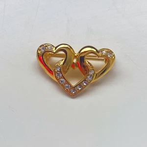 Guldfärgad brosch med hjärtan - Elegant guldfärgad brosch med två sammanlänkade hjärtan dekorerade med gnistrande stenar. Perfekt för att ge en romantisk touch till din outfit. Broschen är cirka 3 cm bred.