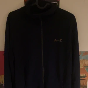 Svart jacka från A-Z - Säljer en stilren svart jacka från A-Z med dragkedja framtill. Jackan har en minimalistisk design med A-Z-logga på bröstet. Perfekt för en casual look.