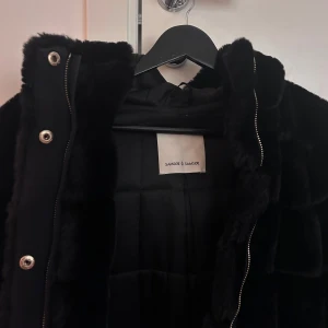 Svart pälsjacka från Samsøe & Samsøe - Snygg svart pälsjacka från Samsøe & Samsøe med dragkedja och knappar. Jackan har en fluffig look och är perfekt för kyligare dagar. Den har långa ärmar och en mysig huva.