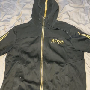 Hugo boss hoodie/S - Snygg svart hoodie från Hugo Boss med gulddetaljer och dragkedja. Den har en huva och ribbade detaljer vid axlarna. Perfekt för en stilren look. Storlek S