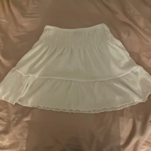 Vit kjol från Gina Tricot - Säljer en söt vit kjol från Gina Tricot i storlek S. Kjolen har en volangdetalj och är perfekt för varma dagar. Den elastiska midjan ger en bekväm passform. Passar utmärkt till sommarens alla tillfällen!