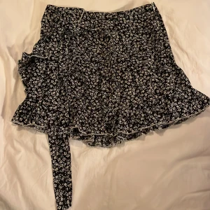 Svart blommig omlottkjol - Charmig svart omlottkjol med vita blommor och volangdetaljer. Kjolen har en knytfunktion i midjan och är perfekt för en somrig look.