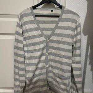 Grå- och vitrandig kofta - Säljer en stilren grå- och vitrandig kofta med v-ringning och knappar framtill. Koftan har långa ärmar och en liten broderad detalj på bröstet. Perfekt för en avslappnad look.