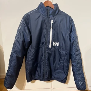Blå vindjacka från Helly Hansen - Snygg blå vindjacka från Helly Hansen med dragkedja och hög krage. Jackan har en praktisk bröstficka med dragkedja och Helly Hansen-logga på bröstet och ryggen. Perfekt för blåsiga dagar.
