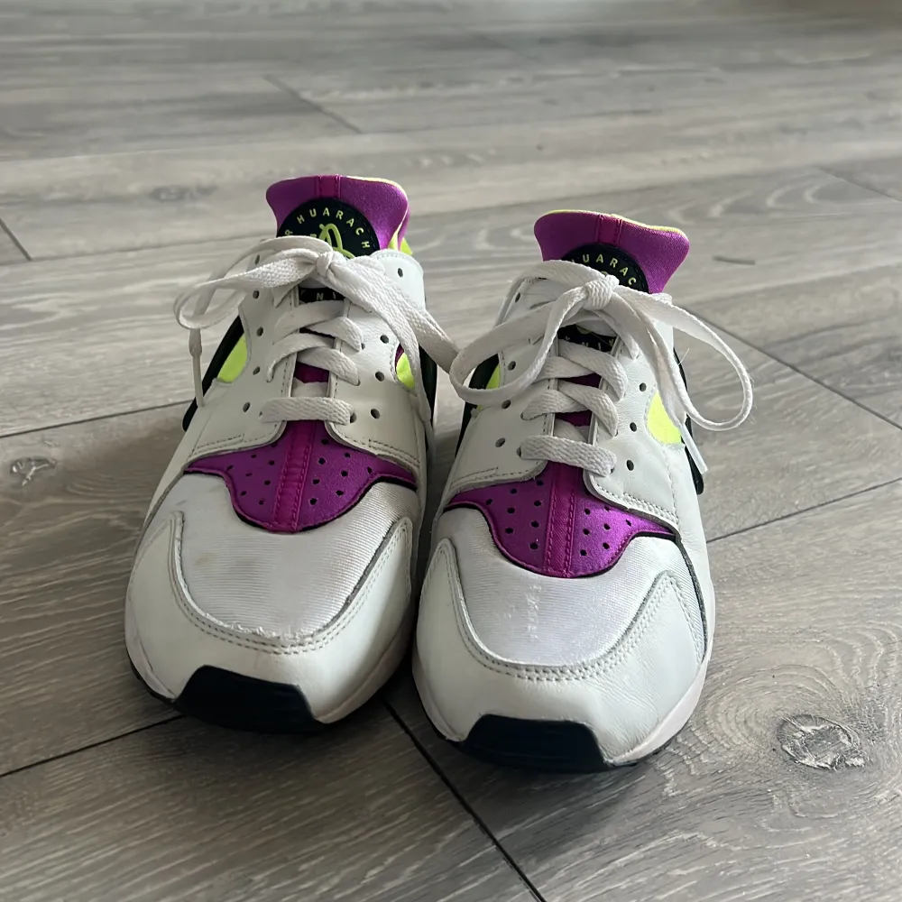 Snygga Nike Air Huarache sneakers i vitt med lila och neongula detaljer. Skorna har en unik design med snörning och en stabil sula för extra komfort. Perfekta för en sportig look.. Kengät.