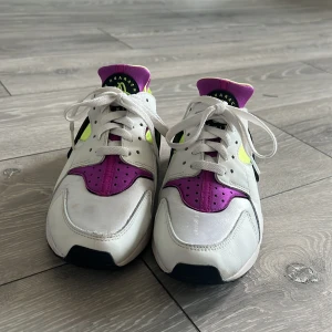 Nike Air Huarache sneakers i vitt och lila - Snygga Nike Air Huarache sneakers i vitt med lila och neongula detaljer. Skorna har en unik design med snörning och en stabil sula för extra komfort. Perfekta för en sportig look.