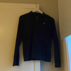Navy blue tröja med quarterzip från Lyle & Scott - Snygg mörkblå tröja från Lyle & Scott med en halv dragkedja och deras ikoniska gula logotyp på bröstet. Perfekt för en stilren och avslappnad look.