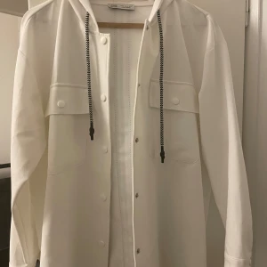 Vit kofta/ hoodie med huva - Snygg vit kofta med huva och svarta snören. Koftan har knappar framtill och två stora fickor på bröstet. Lätt och tunn. Använts 2 gånger och är fortfarande i väldigt bra skick. Strl L MEN passar S och M ( köpt utomlands därav är storleken annorlunda)