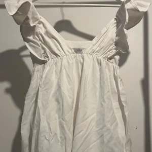 Vit klänning från Chiquelle - Charmig vit klänning från Chiquelle med volangdetaljer på axelbanden. Perfekt för sommarens varma dagar. Klänningen har en luftig passform och är tillverkad i ett lätt material. Perfekt studentklänning 